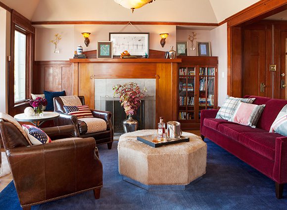 A gentlemanly living room, showing blue carpet, dark brown leather club chairs dressed with American flag pillows, a large modern faux horsehair octagonal footrest used as a table, with whiskey and ice bucket on a tray. On the other side of the footrest is a red couch with several throw pillows. The rear wall has bookshelves either side of a wood fireplace surround and mantel, dressed with artwork. In front of the mantel is a flower arrangement. Light streams in from the windows on the left. Wood paneling surrounds the windows, doors, and acts as cornicing