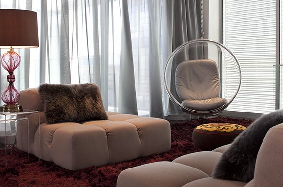 A retro-futuristic living room for a bachelor, with two pink B&B Italia multi-directional sofas with fuzzy throw pillows on a deep red wine-colored shag rug. An Eero Aarnio bubble swing chair hangs just in front of a large set of windows, with a patterned footstool in front. to the left of one sofa sits a clear perspex table with a burgundy table lamp.