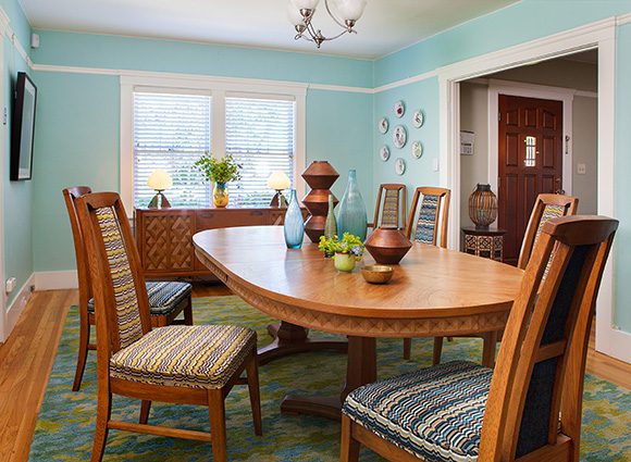 This dining room has light aqua-blue walls with white trim, orangey hardwood flooring and a green-and-turquoise-blue patterned rug. The vintage mid-century modern dining table and chairs are the same orangey tone as the flooring, ranging to a little darker, and the chairs are upholstered with multicolored striped fabrics on seat and center of back. The table is set with several geometric-shaped vases and bowls. A sideboard sits in front of a window at the back. On the wall to the right hang some collectible plates, near the open doorway through to the entrance hall, showing the front door in a dark wood and white trim.
