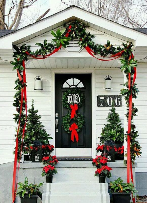 Photo of a residential front entrance with a black front door and grey-and-white painted concrete steps. Swags of fresh greenery are tied with wide red ribbons over the porch-front on a white clapboard surround and hang all the way down to the ground. A pair of matching green wreaths with wide, red bows hang vertically in the center of the black door, adorned with the family's initials. Red poinsettia plants line the top two steps either side, and continue up onto the porch, where they sit in front of 2 taller planters filled with small pine trees and more greenery. Two white front porch lights trimmed with black are hung either side of the door, which has silver door furniture and matching kickplate. The house number on the left side is a large, horizontal rectangular black background with huge numbers cut out, revealing the white clapboard siding behind.