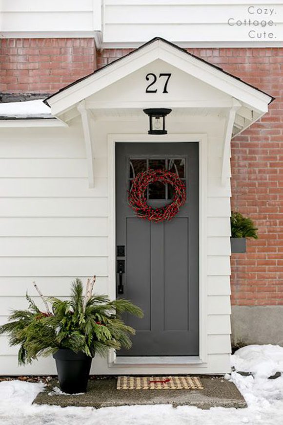 Photo of a small, white clapboard peaked cottage front entrance with a dark charcoal-grey door, adorned with a twisted wood wreath filled with red berries as an understated holiday decoration. To the left of the door is a black pot overflowing with evergreens and white ash sticks. The front porch stone slab, surrounded by white snow, offers a grey-and-cream plaid doormat with a reindeer in red. A peek of red brick topped with white clapboard from the rest of the house shows above and to the right of the white clapboard entrance.