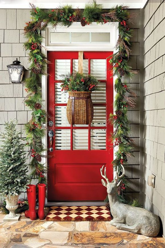 Photo of a bright red front door, decorated with a woven basket containing green cuttings and red berries, hanging in front of the partially open white slatted blinds behind 12 equal-sized front door windows. To the left of the door, on the vertical surface, is a black metal and glass porchlight over sage-green ship-lap cladding. A small pine tree sits in a white grecian urn planter at the far left of the image. A large garland of loose evergreen swags punctuated with red berries and pinecones surrounds the door frame. A pair of red rubber rainboots wait to the left of the brown-and-cream diamond porch mat, sitting atop marbled and striated sandstone pavers in brown, tan, cream, and coppery colors. A concrete grey statue of a seated reindeer or stag with full antlers is nested to the right of the door.