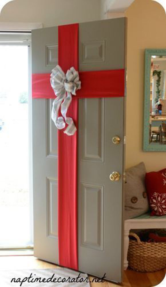 Photo of a sage-green apartment door, opened inward to reveal a Christmas gift decoration of two large, red ribbons crossing with a huge, white fluffy bow in the center, and curled trailing ends hanging down. To the right and behind the door are a seat filled with pillows, a woven basket tucked underneath, and a mirror trimmed in turquoise blue-green.