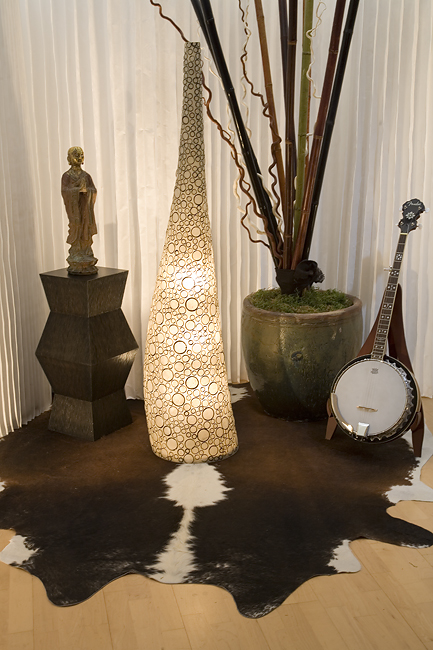 A tall tapering lighted lamp-type shape, with swirling circle patterns. Nearby, a carved statue of a Buddhist monk stands on top of a side table. To the right, a large container holds sticks and vines. All sit atop a cow hide rug over a light wood floor.