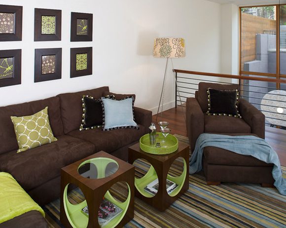 Hillside lounge area includes a brown sofa with throw pillows in lime green, sky blue, and black, a matching striped rug, 2 moveable tables in brown and green, and a matching chair and ottoman with a black throw pillow and sky blue blanket thrown over the ottoman. A standing lamp is placed between the sofa and chair. 6 frames on the wall show artwork, while natural light streams in from the double-height window at the right.