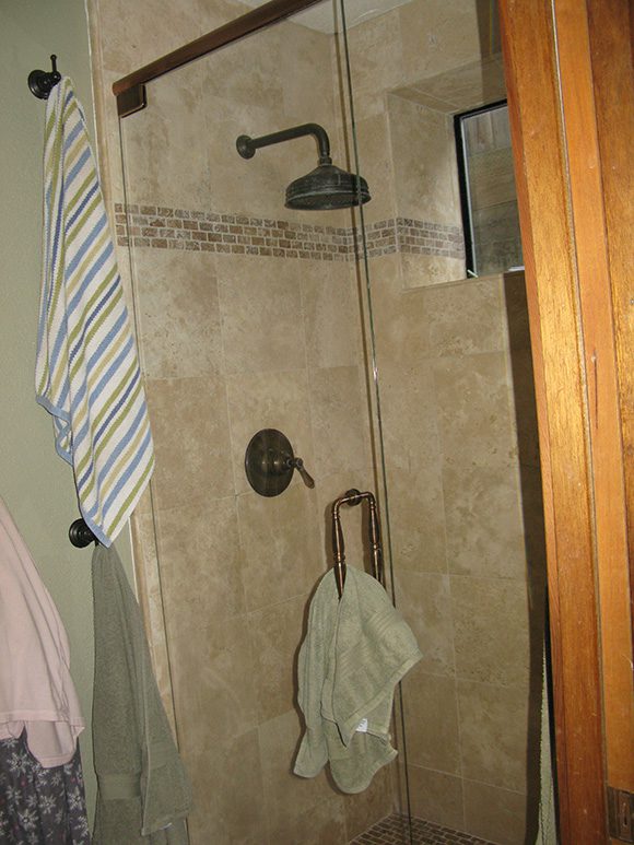 Another 'before' image showing mismatched towels hung from a multitude of hooks, which encourages that mildewy smell in a bathroom. The olde-world-look, faux marble tiles in a creamy tan-gold with white veining, in combination with heavy black fixtures such as the showerhead and handle, say "Outdated bathroom." Even the shower door handle is being used ineffectively as a towel rail. A tiny slice of a high window peeks out at the back of the shower, outlined in black metal framing, and that sickly greeny-grey wall paint color next to the shower does not help the look. Orangey-colored wood shows just at the right of the image, which seems completely out of place.