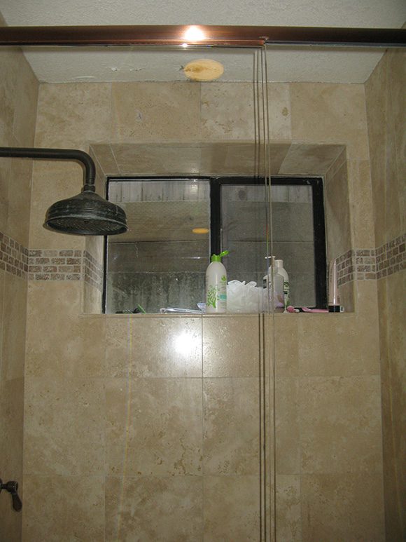 A heavy black 'rainhead'-type showerhead is spoiled by hard-water crusting in this homeowner's "before" image. Faux marbled tiles in tan, gold and white veining don't look classic, they look outdated. Personal items and bottles line the windowsill, illustrating there isn't any storage. The black metal-framed window itself at the back of the shower is still a useful feature and will be kept, but refinished. The mini-brick-like row of accent tiles running around the shower at head-height does nothing to modernize the space.