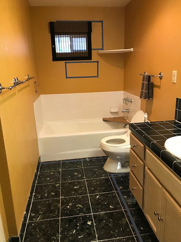 View into an outdated bathroom, with orangey-gold wall color paint, black flooring with white square grouting and reflective flecks, and yellow-gold toned cabinetry with a black countertop surface. There is a small window above the tub, which has unattractive security bars, and a toilet sits between the tub and the counter unit. Multiple towel rails line the walls.