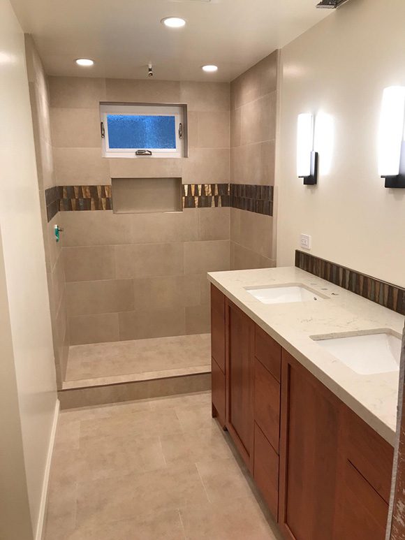 View into a partially-finished light-colored contemporary bathroom. The flooring and step-in shower wall tiles are varied shades of tan, with a center horizontal accent double-line of darker brown and orangey reflective tiles with a triangular shape embedded. Another row of those tiles adorns the back edge of the countertop and sink, but there are no faucet fixtures yet, and no mirrors. The cabinetry is a warm orangey-red that tones with the accent tiling. Wall paint is eggshell tan in color, with vertical rectangular modern wall sconces above the sinks. There are 3 recessed lights in the shower space, and a horizontal rectangular operable privacy window for natural light and fresh air, but no shower doors are installed yet. The toilet has been relocated to another part of the space, outside of this view.