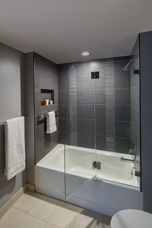 View of a modern bathroom. Flooring is a light tan with square grey grout. Wall color is grey, and the ceiling is white. The tub and toilet are also white. The main shower subway tiling is dark grayish brown, with two single vertical rows of more reflective tiles. The tub and shower are contained by a glass panel and glass door, which hinges above the tub, rather than a shower curtain. A recessed niche allows space for shower and shaving products, while a towel rail is placed on the inside of the shower, and another on the outside. A single recessed light fixture provides illumination.