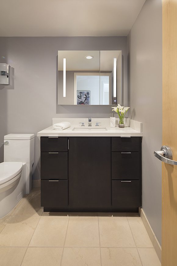 Contemporary bathroom with dark wood cabinetry and silver horizontal drawer pulls, under a white counter surface. Mirrored medicine cabinet contains built-in side lights. The faucet fixture is square and silver with two taps. The toilet is white, sitting to the left of the counter space, which is styled with fresh white lilies and a white and silver soap bottle, with a white towel folded neatly to the left of the sink. The wall color is grey, with a white glass and silver accented rectangular wall sconce, and a single recessed light visible in the mirror.