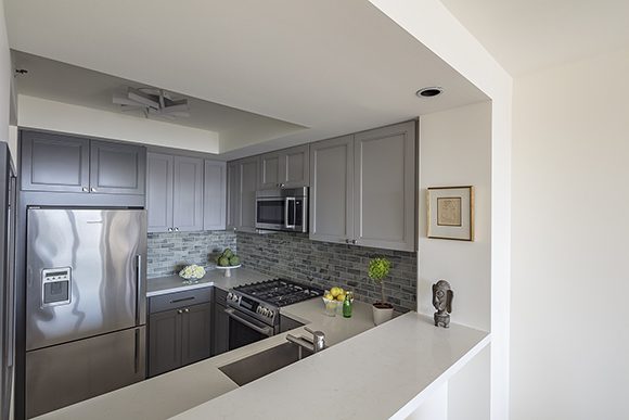 Looking into a kitchen through a dining room pass-through space. At the far left is a large silver-finish fridge and freezer unit, with grey cabinetry above, that continues around the rest of that wall and the right wall, where you see a microwave above and a range and oven unit below. Subway tiles in varied shades of green and grey counterpoint the green plant and fresh fruit displays. Light white-colored worktops are continued in color on the passthrough, which doubles as a dining counter surface. White walls and ceiling continue throughout the rest of the space.