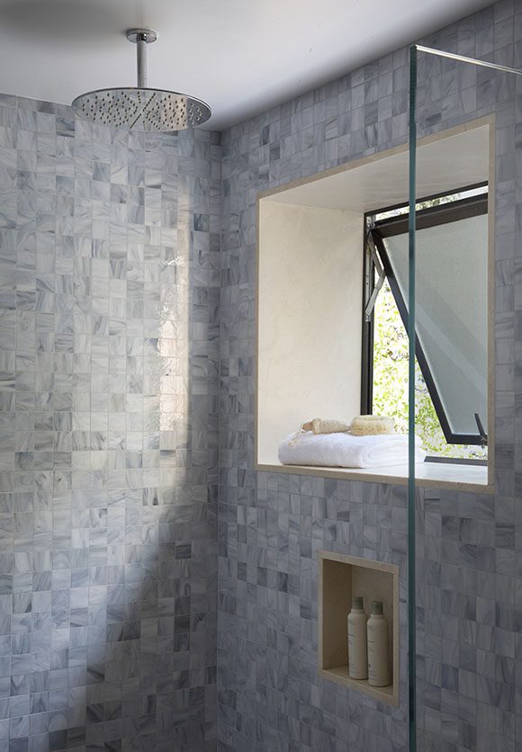 A generous rainhead showerhead is placed near an operable window, dressed with towels and loofahs on the windowsill, while an inset niche creates space for personal item storage at just the right height. The whole space has a light, soft feeling created by mosaic tiles in multi-color hues of blue, grey, and white.