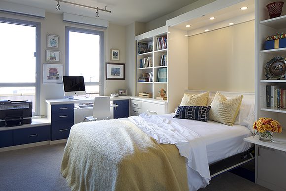 An “after” view of the home office, showing an unmade bed with crisp white linens, yellow blanket, and navy blue accent pillows, in front of the desk and counterspace for an Apple computer, with matching navy blue storage drawers that balance the blue of the wall-bed cabinet, when visible.