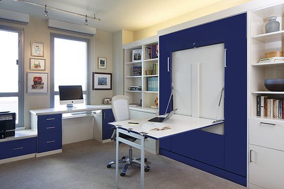 An “after” view of the home office, showing the navy blue custom cabinetry that hides the convertible wall-bed, with the white wood top and grey metal-legged desk folded down, giving additional space for laying out papers, drawing, reading, or computing.