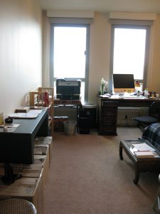 "Before" photo of the home office showing large, tall windows, with all walls lined with boxes, desks and chairs, 2 computers and a printer. Walls are blankly stark.