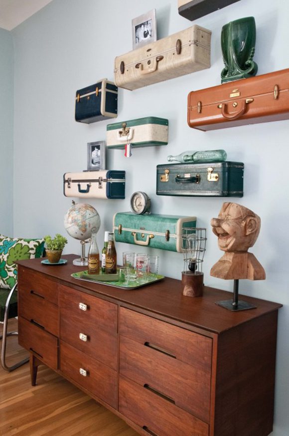 A sleek dark teak or redwood mid-century modern dresser is adorned with sculpture and a globe, while the walls above feature the ends of vintage suitcases hung as shelving and dressed with sculptures, clocks, and art.