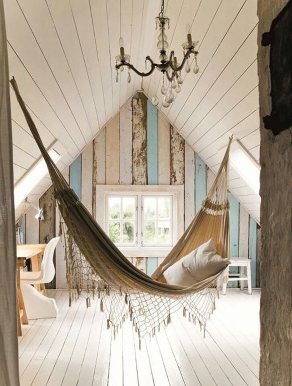 Whitewashed peaked ceiling and floorboards of an attic with a hammock hung in the center. Netting hangs from the bottom of the hammock, while a pillow beckons. Partially stripped, aged painted wallboards adorn the far wall in eggshell blue, off-white with rust, and faded pink, with 2 windows at the center and a desk off to the left side. Above the hammock is hung a small chandelier for that shabby-chic look.