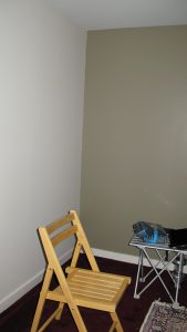 A blank corner with cream wall on left, sage green on right, with white baseboards and red shag carpet. A wooden folding chair sits in the foreground.