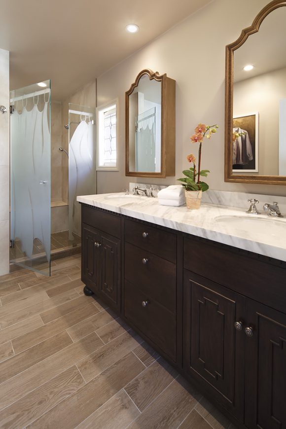 A modern glass shower with curb-less door opens onto multi-toned tan porcelain tiling on the floor, pure white railroad tiles on the bottom half of shower walls, and hexagonal grey and white tiles on the upper half. Dark wood cabinetry contrasts with the white marbled countertop, and a pair of mirrors over his-n-her sinks provides plenty of space for getting ready together.