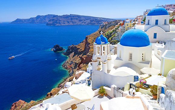 View over the romantic whitewashed buildings and blue roofs matching the deep blue water of Santorini, Greece, lapping around the coastline in jewelled greens.