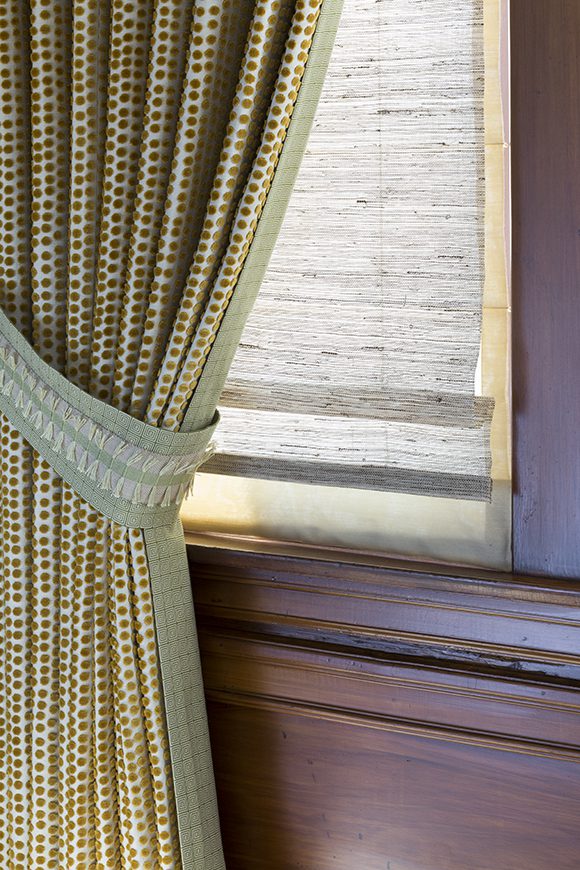 Detail of the window dressings, featuring gold dot-patterned fabric finished with a square green edge pattern and a matching tieback at either end of medium-toned wooden window frames. The windows are covered by textural grass Roman shades, allowing filtered light into the room.
