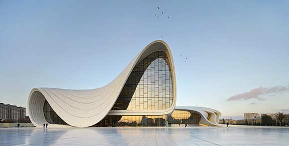 A sexy, curvilicious shaped building rests lightly on a ground of shiny stones, outlined against a soft blue sky. The building itself is white, with black accent lines showing off the curves and hidden surfaces of the windows.