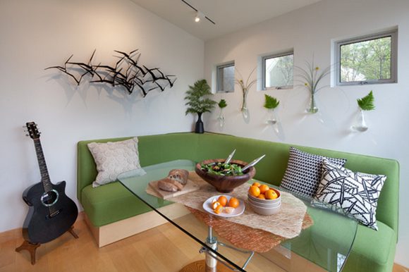Another view of this oddly-shaped corner space, with a lime-green banquette sofa curving around a clear glass pedestal table dressed with salad, fruit, and bread for a snack. The rear wall has 3 windows, accented by 5 small wall vases filled with a single branch of greenery each. On the left wall is a metal relief art piece of birds in a flock. Standing to the left of the banquette is a black guitar on a small wooden stand.
