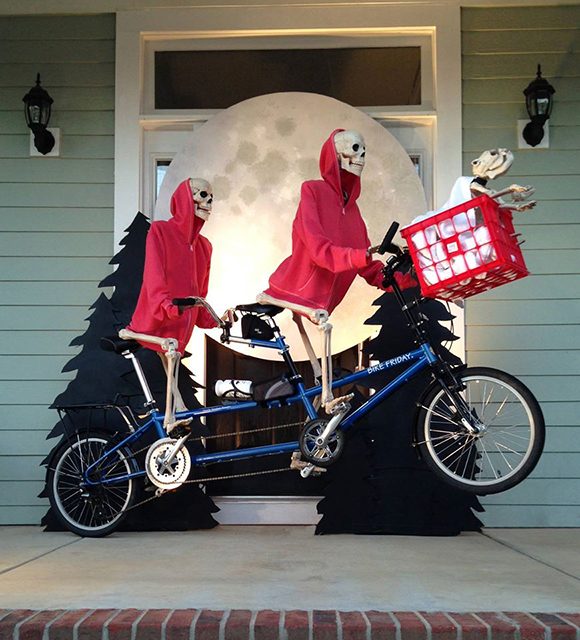 Photo of a front porch with a double bicycle ridden by two skeletons in red hooded sweatshirts and a bike basket with a small dog skeleton, suspended above the porch decking, in front of evergreen trees and a large moon