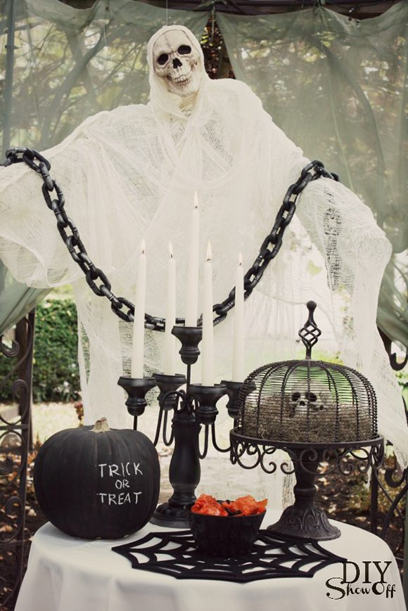 Photo of a skeleton swathed in white gauze with a single black chain across its arms, hanging down in front. On the white cloth dining table in the foreground, a black candelabra with white candles, a black pumpkin emblazoned with Trick or Treat, a wire metal cake stand with a small skull on a bed of moss, and a bowl of orange-wrapped candies on a spiderweb table mat are ready for neighborhood visitors to snack on.