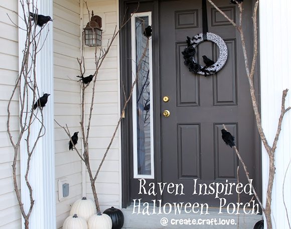 In a subtly designed Halloween decorated entrance photo, the basic light-colored front porch and black front door have a vertical left window featuring a painted white highlight wood surround. The porch is decorated with black ravens sitting atop sticks that look like bare-branched trees. All black and all white pumpkins surround the bases of the trees. On the door, a handmade black-and-white wreath hangs from a black ribbon, adorned with a black bird silhouette, holding a black branch.