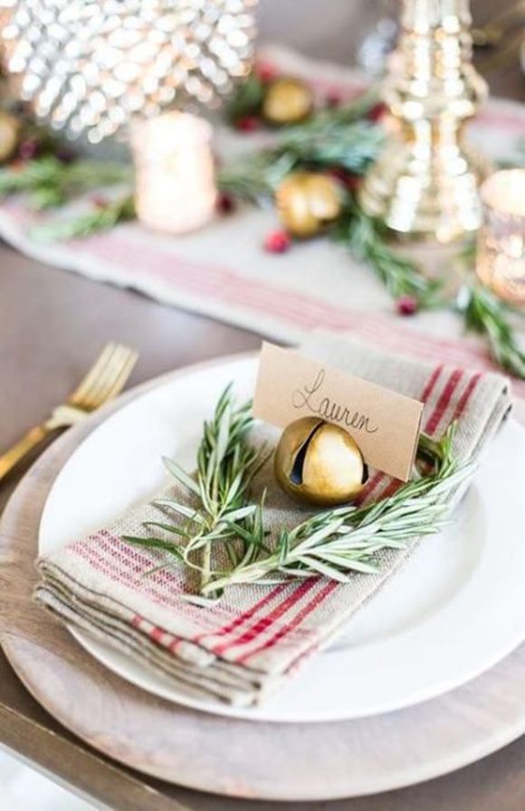 A beautiful country table setting with a red striped linen napkin, sprig of evergreen, and a large sleigh bell to hold the nametag, reading Lauren in script. The white plate is surrounded by gold tableware, gold candlestick holder, and silver and gold candles accented with more evergreen trimmings and red berries, out of focus in the background.