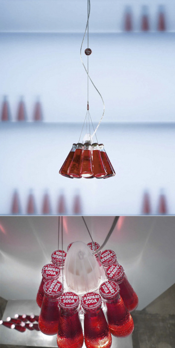 Two images, one longer one above the other. The top image shows a light fixture on a single wire with the power cable snaking around it. Ten Campari red soda bottles are suspended by multiple wires attached to the central cable. In the bottom image, a top-down view of the light fixture allows you to see the bottle caps saying Campari Soda, and a clear plastic cone-shaped top piece. Red light is cast upon a white wall behind. Eight additional bottles of soda are on the table below, out of focus.
