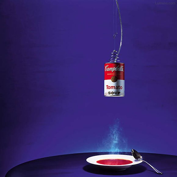 A can of Campbell's Tomato Soup is suspended by a spring above a table where a bowl of piping hot soup and a spoon await, steam rising in the light of the pendant lamp emanating from the bottom of the can. The entire background of the image has a deep blue color. The can of soup is reminiscent of a famous pop-art piece by Andy Warhol.