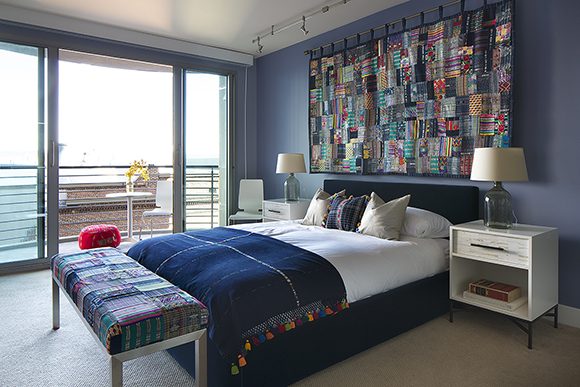 Image of a bedroom with sliding glass doors to the far left, showing a bed with blue velvet headboard, white sheets, and a blue throw pillow with multi-colored tassels flanked by two off-white throw pillows, echoing the multi-colored tassels on the blanket covering the bed. Above the headboard is a quilt made of many multi-colored and textured rectangles of fabric, sewn together. To either side, light-colored bedside tables with a single drawer each hold books underneath in an open space, and on top, glass table lamps are topped by an off-white lampshade. At the foot of the bed, the patchwork quilt material is repeated as custom upholstered fabric.