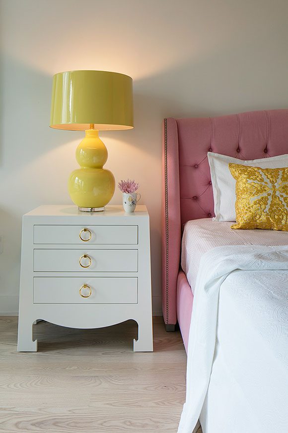 Detail of a feminine bedroom, with light-colored walls and wood flooring. The white bedside table has 3 drawers with gold ring pulls for handles. A bright yellow bubble lamp and shade sits atop the table, to the left of a partial view of a pink upholstered padded headboard with crisp white sheets and a yellow patterned throw pillow.