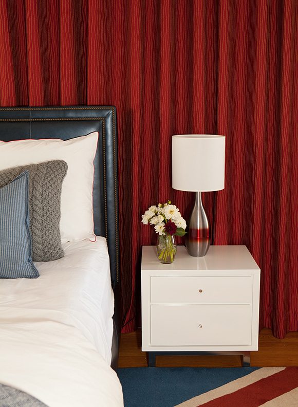 Closeup view of the bedroom. To the left, a dark brown padded upholstered bed with white sheets, layered with white, grey, and blue pillows. A white bedside table, 2 drawers, with a bowling pin lamp and white shade, next to that, a glass vase with fresh flowers. Behind the bed hangs a bright red curtain with wavy blue pinstripes. Warm reddish wood flooring is underneath the Union Jack flag rug.