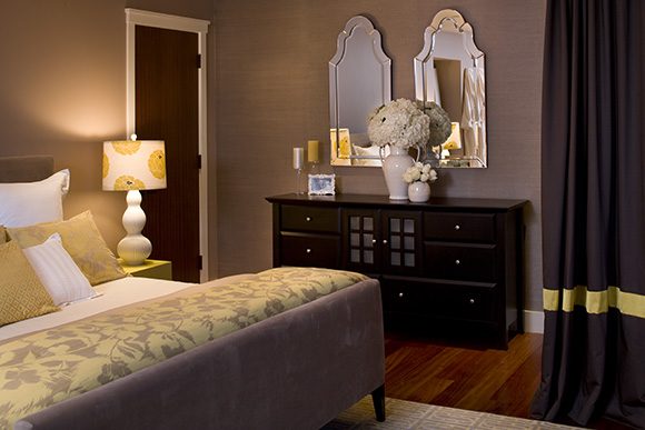 Another view of the luxurious contemporary bedroom. On the right, a dark floor-length curtain with a gold band detail near the bottom. Straight ahead, you see a dark wood dresser, with candles, a framed photo, and two white vases of flowers on top. Above that, hung on the wall, are two rounded-top mirrors, side by side. To the left, a dark brown door with white trim. Further to the left, the bed is upholstered in a grey padded microfiber material, soft to the touch. White pillows behind another layer of yellow patterned pillows, behind another layer of white and yellow and silver patterned throw pillows. A yellow and silver floral bedspread tones with the yellow floral lampshade and white bubble lamp, sitting atop a yellow lacquered bedside table.