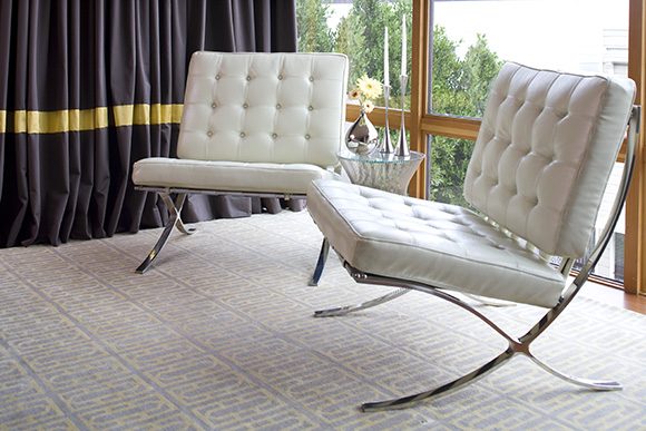 Closeup of two white leather upholstered Barcelona chairs with silver metal frames, sitting atop a grey and yellow geometrically patterned rug. At the back left, you see part of a floor-length dark curtain, with a yellow band of fabric about a foot above the floor.