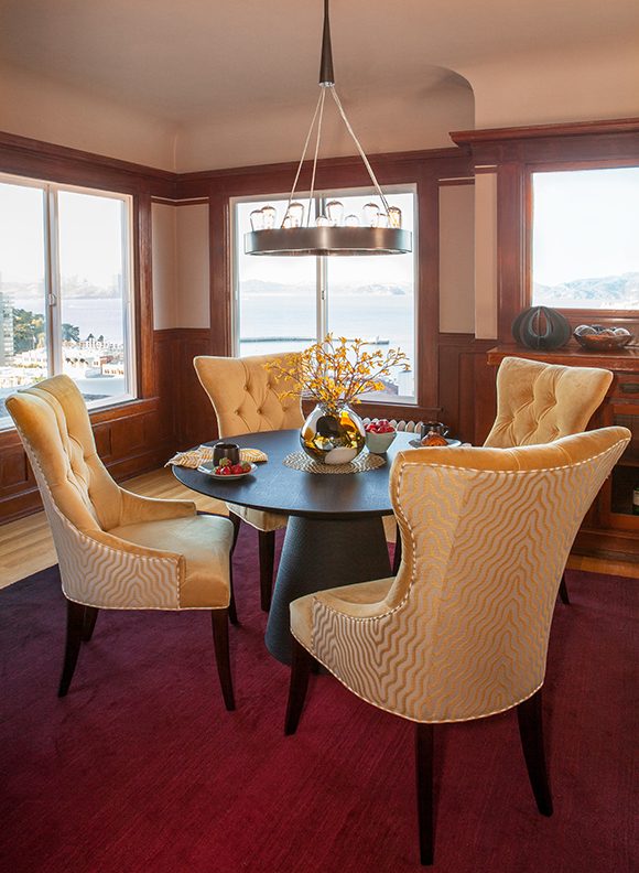 Surrounding a dark wood pedestal dining table, sit four yellow velvet upholstered dining chairs with buttons, and a wavy yellow and gold pattern on the backs. Dark cherry wood flooring is banded by a lighter blond wood at the edges of the room. Behind the dining table is a corner with windows on either side. Above the dining table hangs a round metal pendant light with multiple Edison lightbulbs. On the table are small plates with mugs and fruit, with yellow striped fabric napkins.
