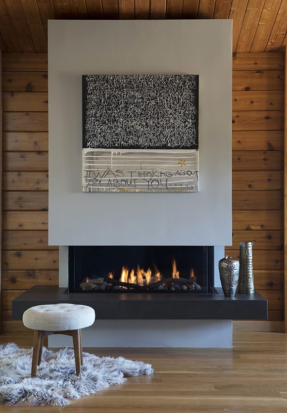 Photo of a contemporary fireplace, surrounded by a knotty cedar wood slatted cabin interior, with tan wood flooring. The fireplace is light grey in finish, without a mantle. Artwork is hung above the fireplace, and two vases sit on the fire surround, finished in black. In front of the fire sits a small grey upholstered round stool with brown wood legs in the Scandanavian style, atop a wooly grey rug.