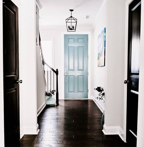 Photo of a hallway with dark wood flooring, light grey-colored walls, white trim, and two black doors either side of the hallway. At the end, the main front door is a robin's egg blue-green.