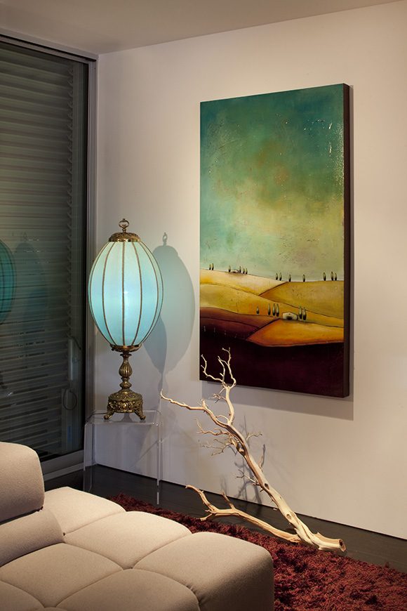 living room corner in a modern bachelor's condo, showing an unusual lamp in blue glass with brass metal fixture, next to a large oil painting of a landscape. In front of the painting on the floor sits a white dried tree branch, which draws your eye down to the red rug in front of a pale pink sofa just visible at the front left of the image.