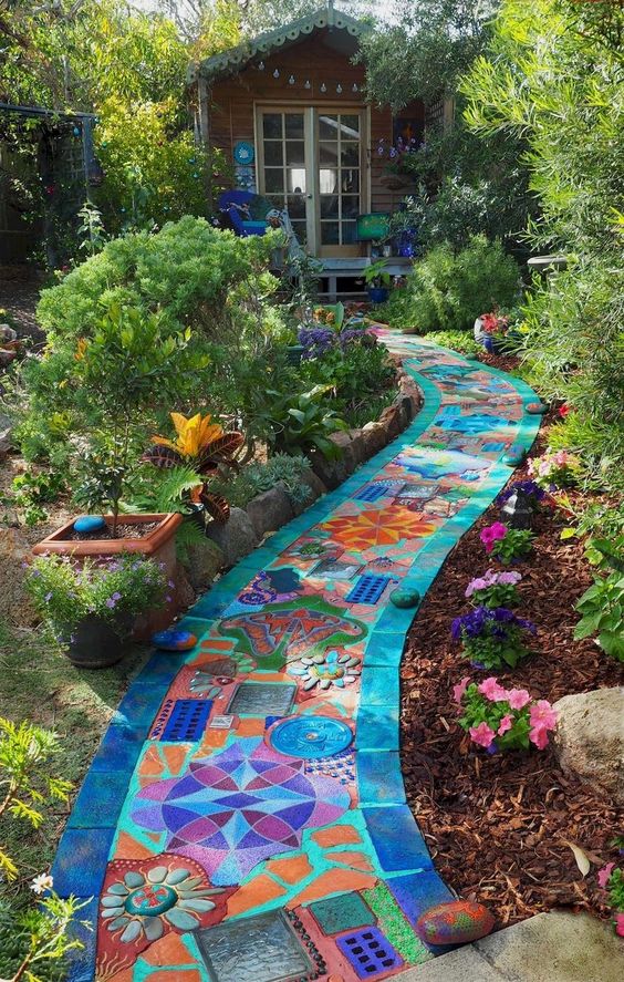 A cobalt-blue bordered pathway of multicolored and patterned tiles stretches through flowers and greenery to a cottage-style shed at the back of a garden.