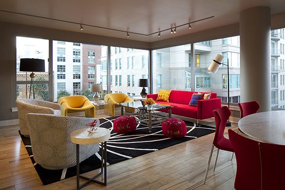 A high-rise condo's living room shows two window walls meeting at a corner with a view of the city at the rear, and track lighting above. In front, a red sofa with yellow and blue throw pillows, a pair of yellow modern chairs, a pair of yellow and grey patterned tub chairs, surround a glass coffee table with two red poufs nearby. All sit atop a large black rug with white radial lines emanating from one end. A dining table peeks from the right side, surrounded by red modern dining chairs.