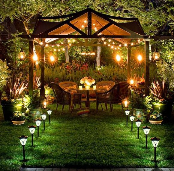 A pathway is created by mid-calf-height lights in a row either side to a pergola. A canopy effect is created with string lights from the pergola's roof interior. Tall stake-lights provide area lighting just behind the seating, while table candles give that perfect intimate touch. Trees and plants are uplit dramatically all around.