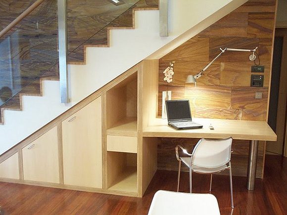 A wooden built-in home office is created under stairs by adding both closed and open storage cubbies, plus a desk, lighting, chair, and bulletin board on the wall. This one uses blonde wood and a white chair.