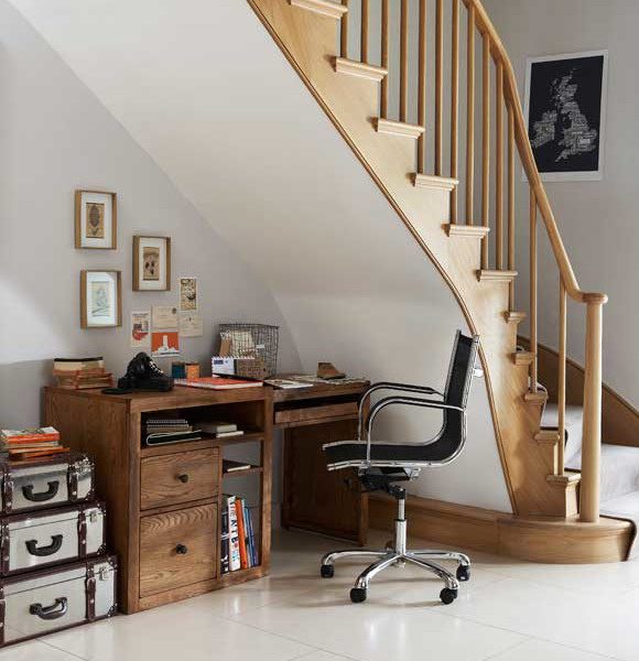 A home office under the stairs, showing a dark wood desk with two drawers, plus a pull-out keyboard surface, and open storage cubbies. A black rolling chair with silver arms makes it easy to get in and out of the space, on a white tiled kitchen floor. Small framed pictures on the wall create a pleasant workspace, while painting the underside of the stairs in white allows light to bounce around the space easily.