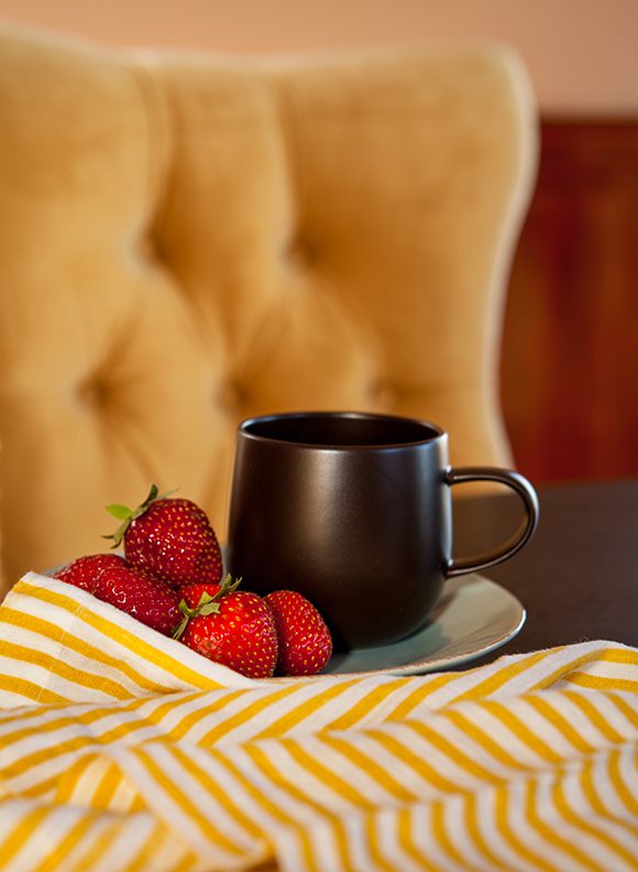 Closeup of a black mug of coffee or tea with four bright red strawberries on a white plate, with a yellow and white striped napkin casually crumpled in front on a dark wood table. Slightly out of focus at the rear of the image is a yellow velvet tufted dining chair back.