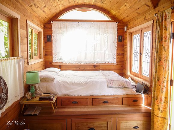The interior of a tiny house by Zyl Vardos, showing an arched window at the back above a large square window covered by a lace curtain. A queen-sized bed viewed from the side is set upon two tiered rows of wooden drawers. The ceiling, walls and drawers are all made of the same warm knotty wood, with four square windows allowing light to pour in from either side.
